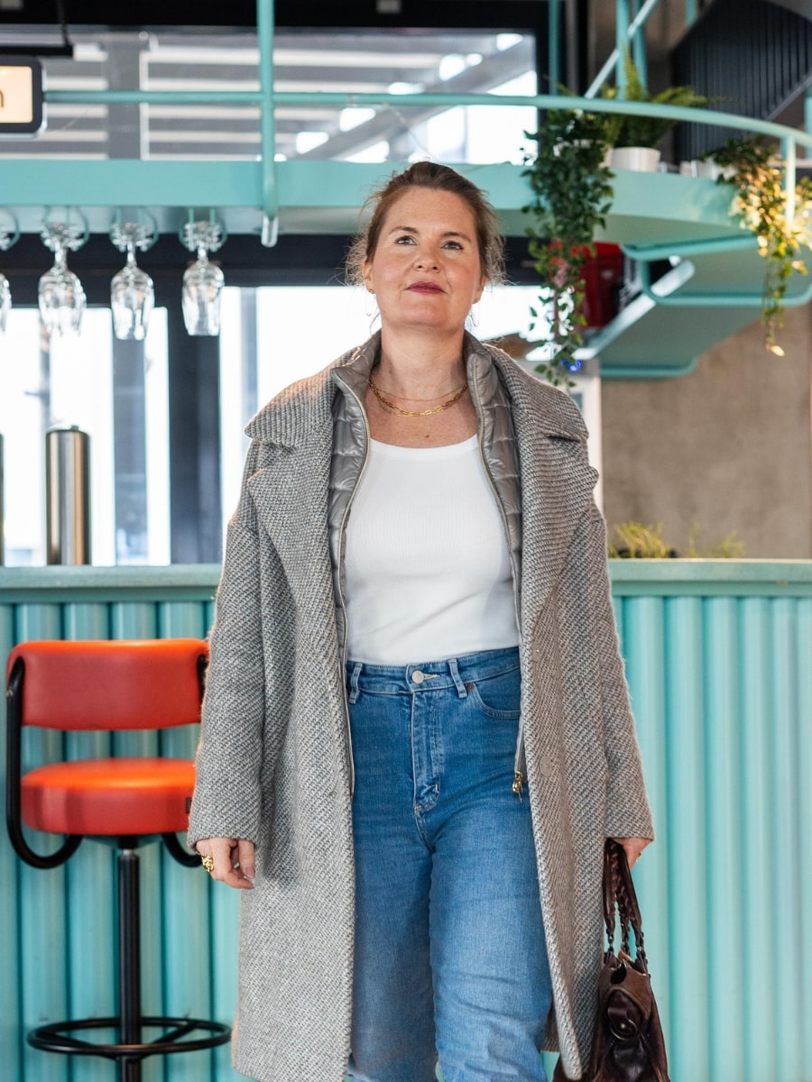 Julia Späth steht in einem modernen Café-Bereich mit hellblauer Theke, trägt grauen Mantel, Jeans und hält eine Handtasche.