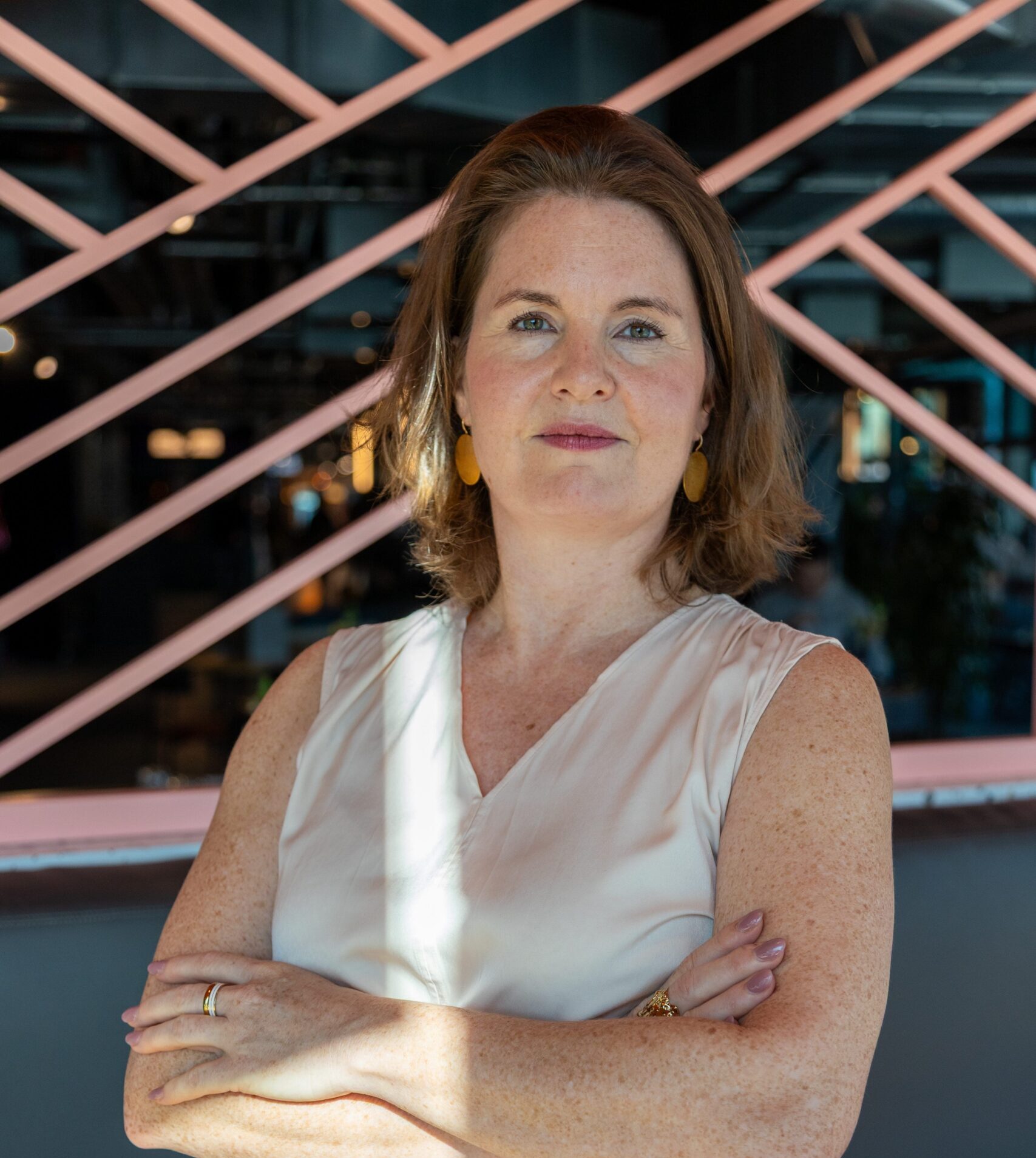 Julia Späth mit verschränkten Armen, selbstbewusster Blick, vor moderner rosa Wand im Café.