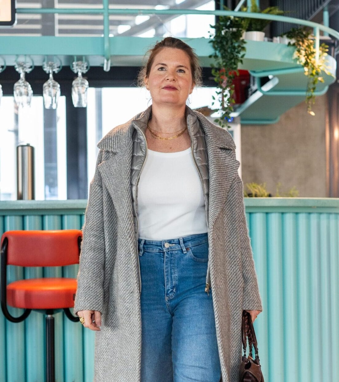 Julia Späth steht in einem modernen Café-Bereich mit hellblauer Theke, trägt grauen Mantel, Jeans und hält eine Handtasche.