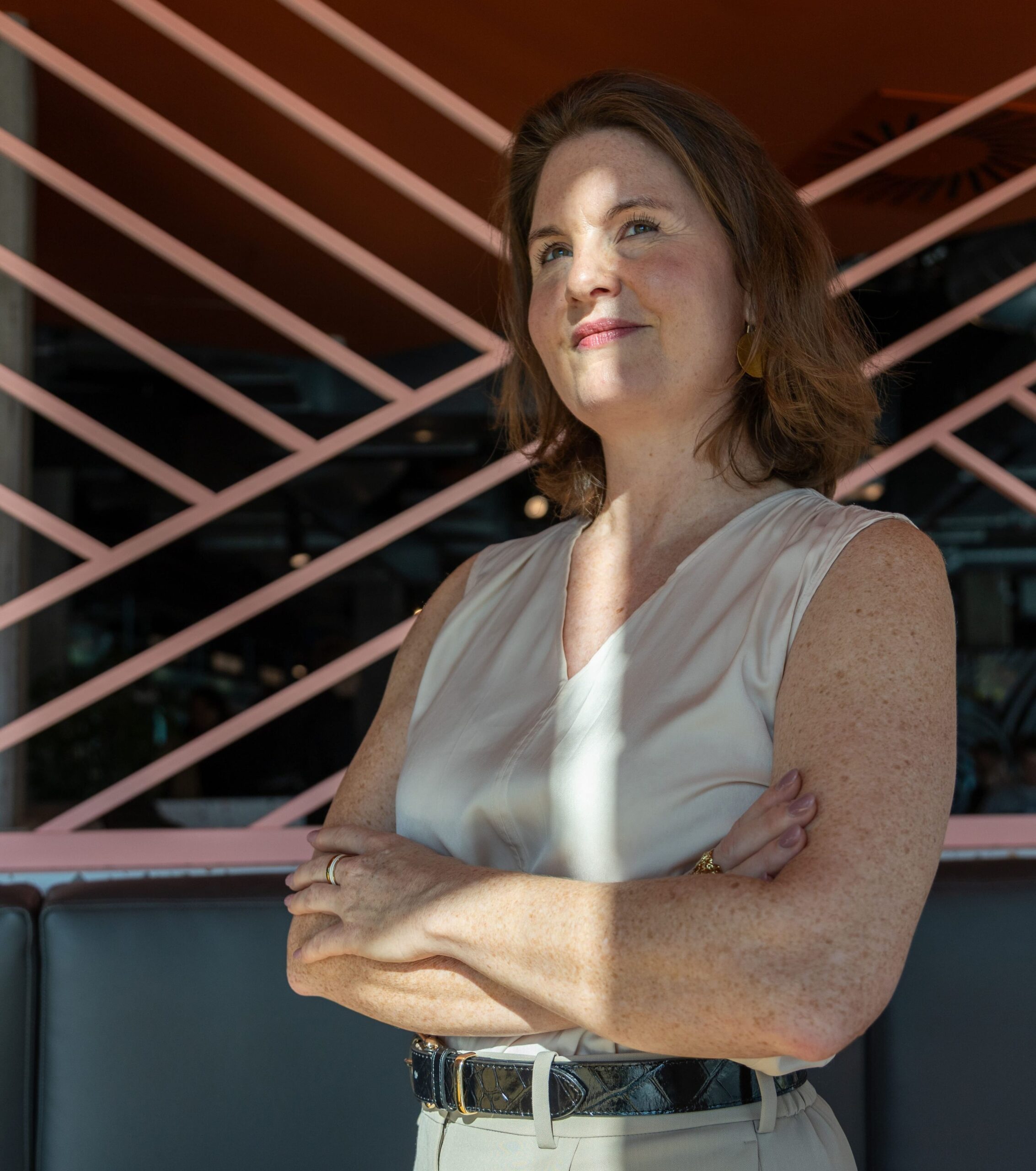 Julia Späth mit verschränkten Armen, selbstbewusster Blick, vor moderner rosa Wand im Café.