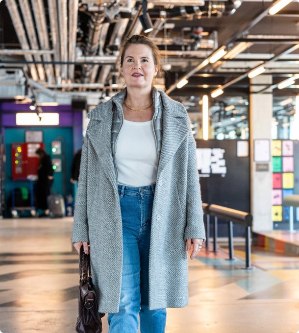 Julia Spaeth läuft durch ein modernes Gebäude mit offenen Decken und bunten Elementen, trägt grauen Mantel