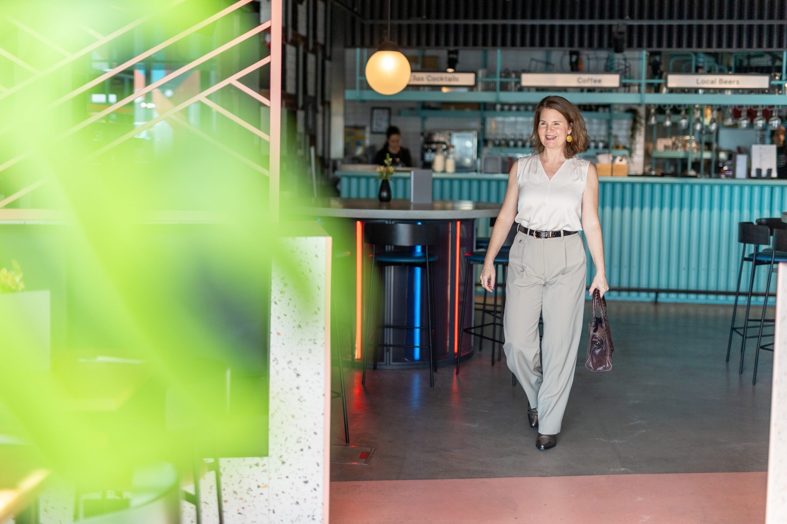 Julia Späth betritt ein stylisches Café mit türkisfarbener Einrichtung, trägt helle Bluse und Hose.
